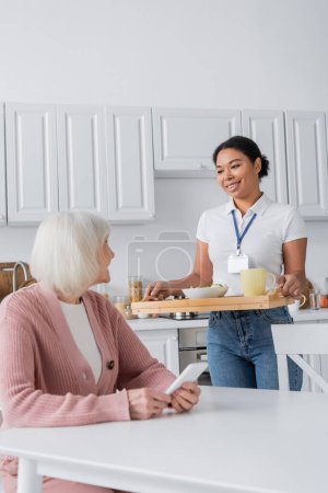 feliz trabajador social multirracial sosteniendo bandeja con almuerzo para mujer mayor con pelo gris 