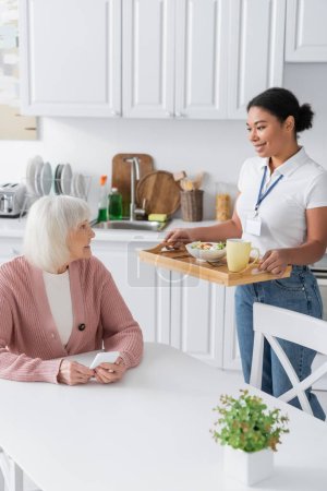 fröhliche multiethnische Sozialarbeiterin hält Tablett mit Mittagessen für Seniorin mit grauen Haaren 