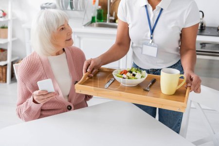 Sozialarbeiterin hält Tablett mit Mittagessen für glückliche Seniorin mit grauen Haaren 