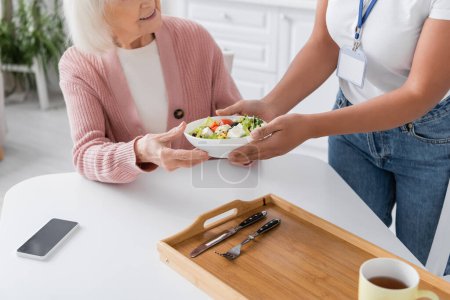 cropped view of multiracial social worker giving bowl with salad to happy senior woman 