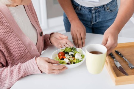 cropped view of multiracial social worker serving lunch near senior woman 
