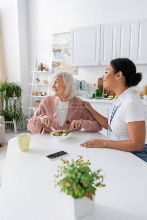 glückliche Seniorin mit grauen Haaren beim Mittagessen neben fröhlicher multiethnischer Sozialarbeiterin 