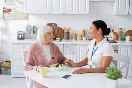 femme à la retraite avec les cheveux gris tenant la main avec soignant multiracial joyeux à côté du déjeuner sur la table 