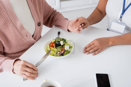 top view of senior woman holding hands with multiracial caregiver next to lunch on table 