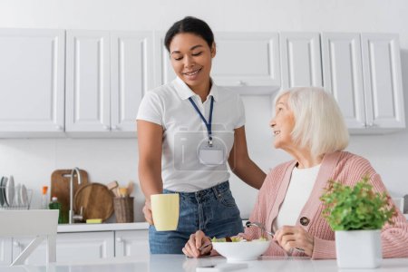 happy multiracial social worker holding cup of tea near retired woman with grey hair having lunch in kitchen 
