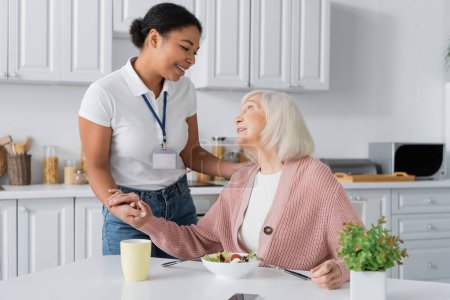 fröhliche multirassische Sozialarbeiterin hält die Hände einer dankbaren Rentnerin 
