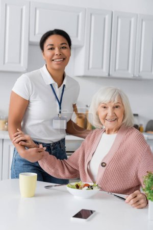 glücklich multirassische Sozialarbeiterin hält Hände einer dankbaren Rentnerin, während sie in die Kamera schaut 