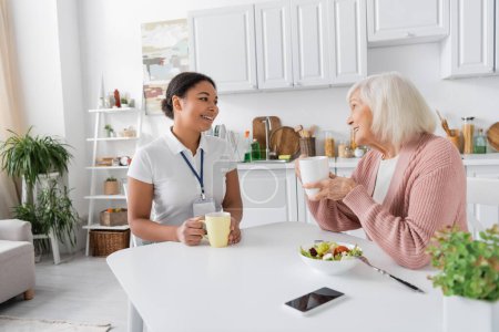 fröhliche multiethnische Sozialarbeiterin beim Tee mit Seniorin in der Küche 