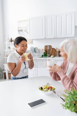 fröhliche multiethnische Sozialarbeiterin beim Tee mit positiver Seniorin in der Küche 
