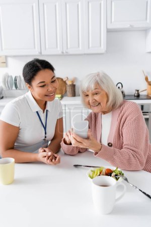 happy senior woman using smartphone near multiracial social worker at home 