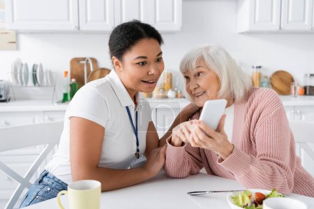 mujer mayor feliz mostrando teléfono inteligente para sorprendido trabajador social multirracial en casa 