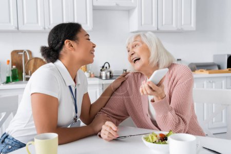 heureuse femme âgée tenant smartphone près joyeux travailleur social multiracial à la maison 