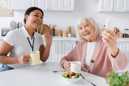 Glückliche Seniorin hält Smartphone in der Hand, während fröhliche multiethnische Sozialarbeiterin während Videoanruf winkt 