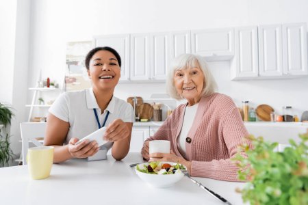 Téléchargez les photos : Heureux travailleur social multiracial tenant smartphone pendant le déjeuner avec une femme à la retraite dans la cuisine - en image libre de droit