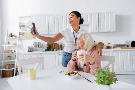 fröhliche multiethnische Sozialarbeiterin macht Selfie mit Rentnerin während des Mittagessens in der Küche 