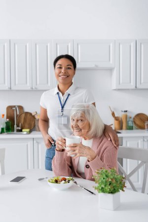 Foto de Feliz trabajador social multirracial sonriendo con una mujer jubilada durante el almuerzo en la cocina - Imagen libre de derechos