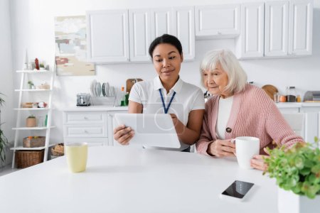 Foto de Feliz trabajador social multirracial sosteniendo tableta digital cerca de la mujer mayor en la cocina - Imagen libre de derechos