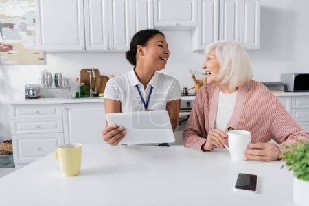 cheerful multiracial social worker holding digital tablet near senior woman in kitchen 