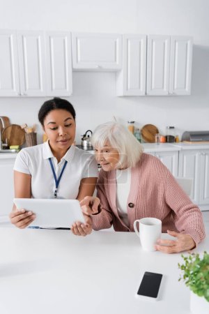 happy multiracial social worker showing digital tablet to senior woman in kitchen 