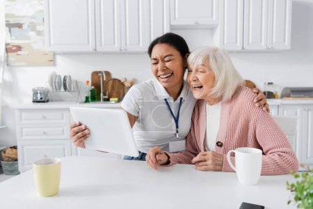 fröhliche multiethnische Sozialarbeiterin hält digitales Tablet in der Hand und lacht mit Seniorin in Küche 