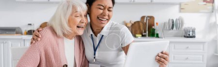 fröhliche multiethnische Sozialarbeiterin hält digitales Tablet in der Hand und lacht mit Seniorin in Küche, Banner 