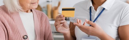 cropped view of multiracial social worker holding credit card near senior woman at home, banner 