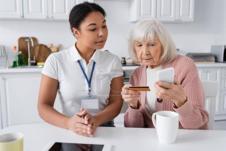 Brünette Sozialarbeiterin schaut Seniorin mit Kreditkarte und Smartphone beim Online-Shopping an 