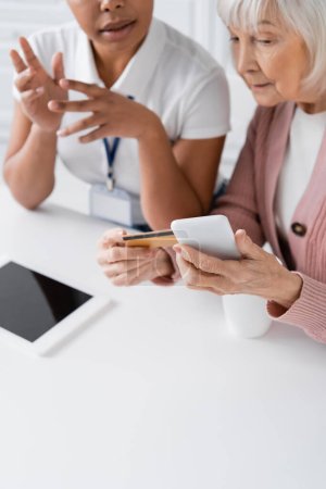 travailleur social multiracial expliquant femme âgée avec carte de crédit et smartphone comment faire des achats en ligne 