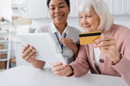brunette multiracial social worker holding digital tablet near happy senior woman with credit card 