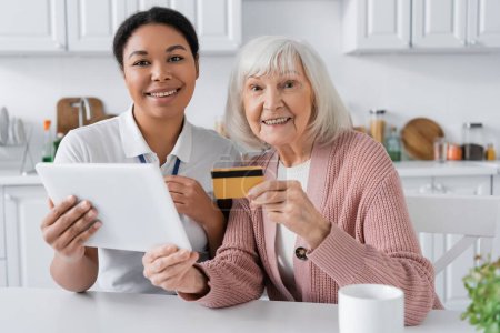 fröhliche multiethnische Sozialarbeiterin hält digitales Tablet in der Nähe positiver Seniorin mit Kreditkarte 