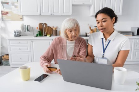 Sozialarbeiterin mit Laptop erklärt Seniorin mit Kreditkarte, wie man online einkauft 
