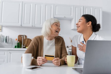 alegre multirracial trabajador social sosteniendo taza cerca feliz senior mujer con tarjeta de crédito y gadgets  