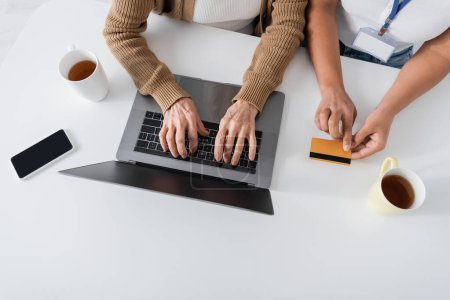 vista superior de la mujer mayor escribiendo en el teclado del ordenador portátil cerca de trabajador social multirracial con tarjeta de crédito 