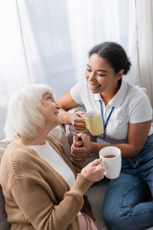 cheerful multiracial social worker chatting with senior woman while having tea in living room 