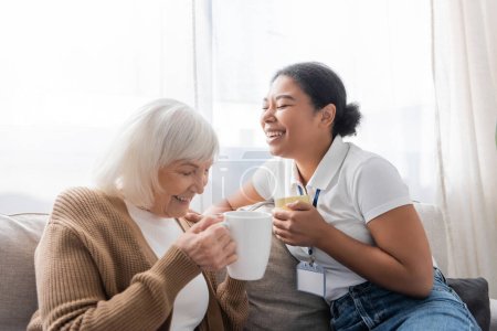 alegre trabajador social multirracial riendo con mujer mayor mientras toma el té en la sala de estar 