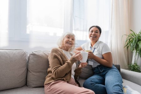 happy multiracial social worker sitting on sofa with senior woman while having tea in living room 