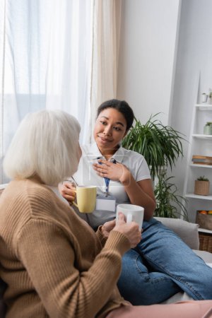 sentimental multiracial social worker chatting with senior woman while having tea in living room 