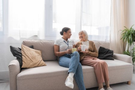 happy multiracial social worker having tea with senior woman in living room 