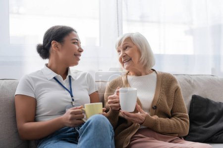 fröhliche multirassische Sozialarbeiterin beim Tee und im Gespräch mit einer Seniorin im Wohnzimmer 