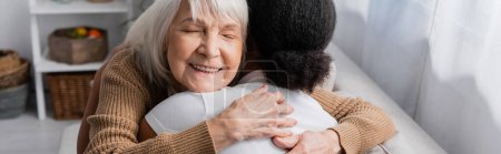 mujer mayor feliz con los ojos cerrados abrazando trabajador social multirracial en la sala de estar, pancarta 
