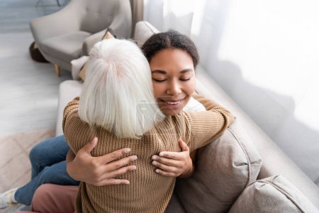 fröhliche multiethnische Sozialarbeiterin mit geschlossenen Augen, die sich mit einer Seniorin im Wohnzimmer umarmt 