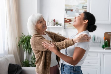 positive multiracial social worker and senior woman laughing and hugging in living room 
