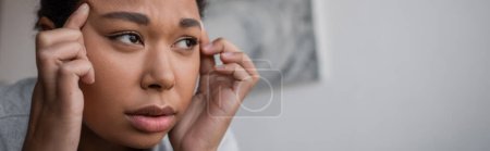 Depressed multiracial woman touching head and looking away at home, banner 