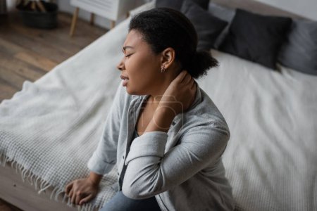 Téléchargez les photos : Vue latérale de la femme multiraciale bouleversée avec dépression touchant le cou tout en étant assis sur le lit - en image libre de droit
