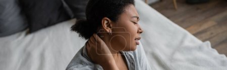 Foto de Vista lateral de la triste mujer multirracial tocando el cuello cerca de la cama en casa, pancarta - Imagen libre de derechos
