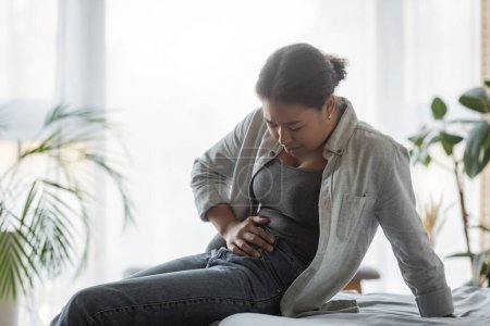 Téléchargez les photos : Jeune femme multiraciale souffrant de douleurs abdominales au lit à la maison - en image libre de droit