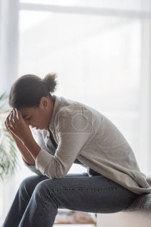 Depressive multiethnische Frau berührt Kopf, während sie zu Hause auf dem Bett sitzt 