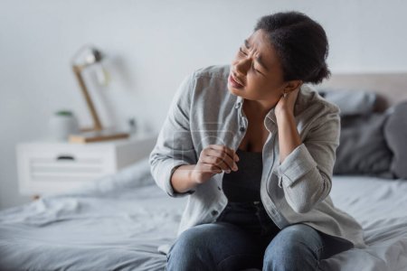 Mujer multirracial cansada tocando el cuello y sufriendo de dolor en el dormitorio en casa 