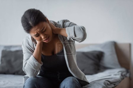 Verspannte multirassische Frau berührt Hals, während sie zu Hause auf dem Bett sitzt 