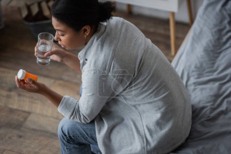 Blick von oben auf traurige multirassische Frau, die Pillen betrachtet und ein Glas Wasser im Schlafzimmer hält 